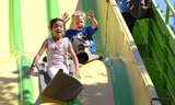 Two children gleefully sliding down a yellow and green slide at the San Mateo County Fair